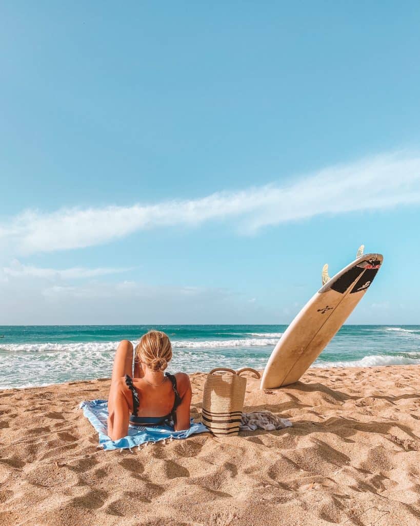 Girl laying on beach watching ocean with surfboard next to her