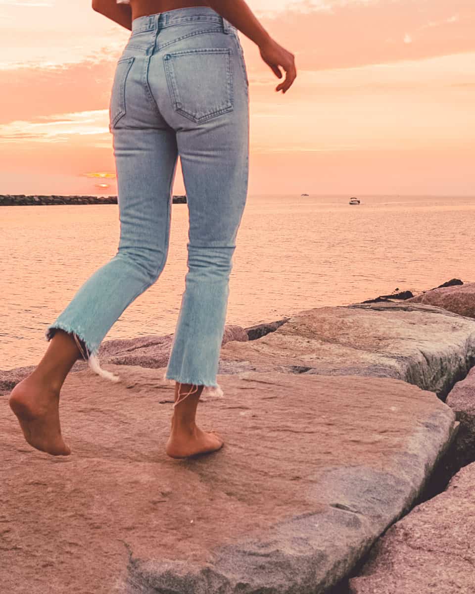 womans legs wearing jeans walking barefoot down a jetty in montauk at sunset
