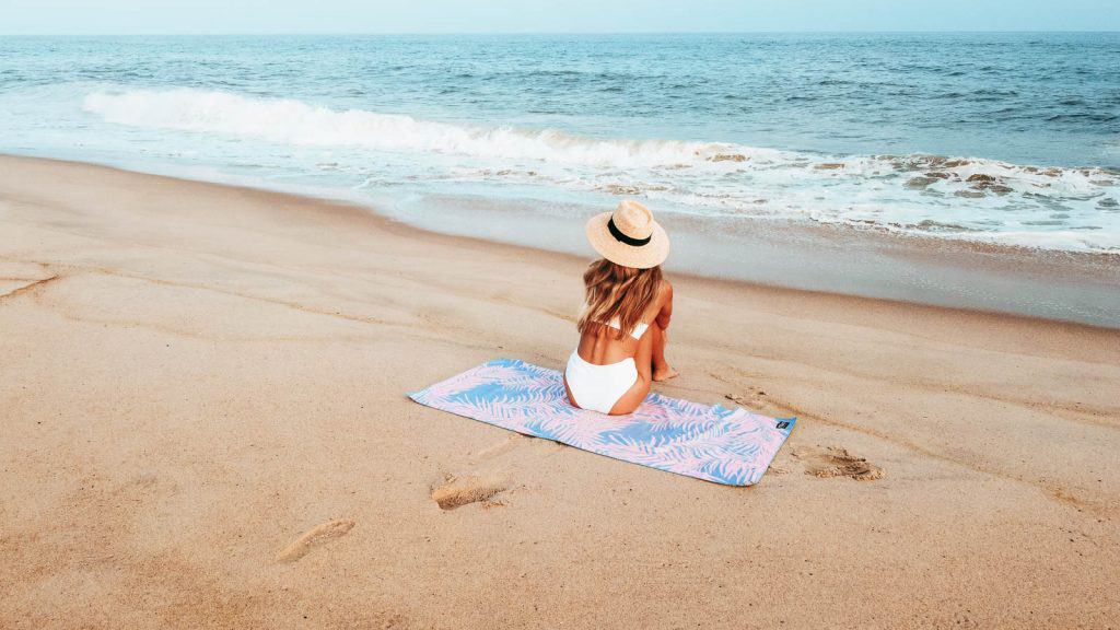 Woman in straw hat sitting on towel in front of the ocean on the sand.