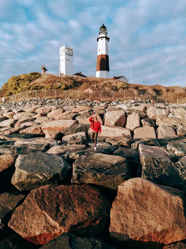 woman in red jacket standing on rock bouulders in front of the montauk lighthouse